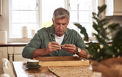 Buy stock photo Cropped shot of a relaxed senior man preparing to roll a marijuana joint to smoke inside the kitchen at home during the day