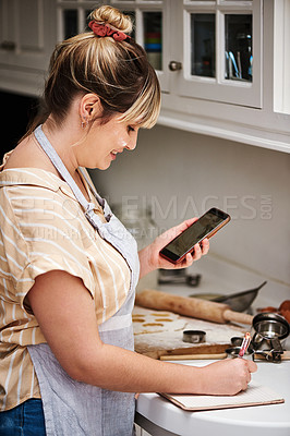 Buy stock photo Cropped shot of a young woman making notes while using her cellphone in her kitchen