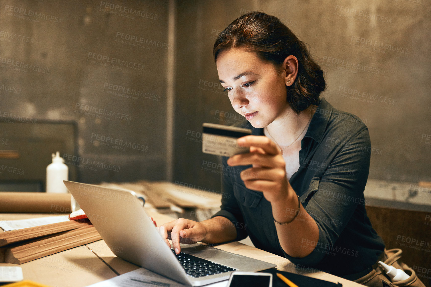 Buy stock photo Cropped shot of an attractive young female carpenter holding her credit card while doing online shopping inside of a workshop at night