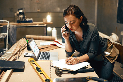 Buy stock photo Cropped shot of an attractive young female carpenter working on her laptop while talking on her cellphone inside of a workshop