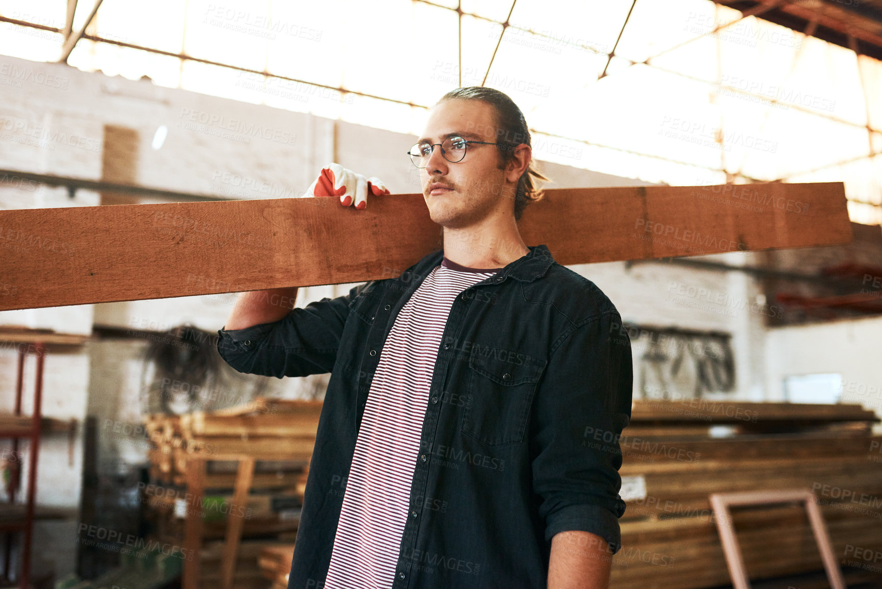 Buy stock photo Shot of a handsome young carpenter carrying a wooden plank over his shoulders inside a workshop