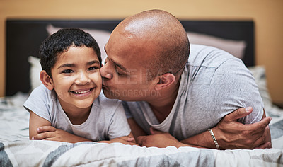 Buy stock photo Portrait of a cheerful little boy receiving a kiss on his cheek by his father while they hang out on the bed at home during the day