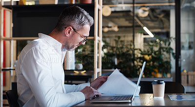 Buy stock photo Shot of a mature businessman using a laptop and going through paperwork in a modern office