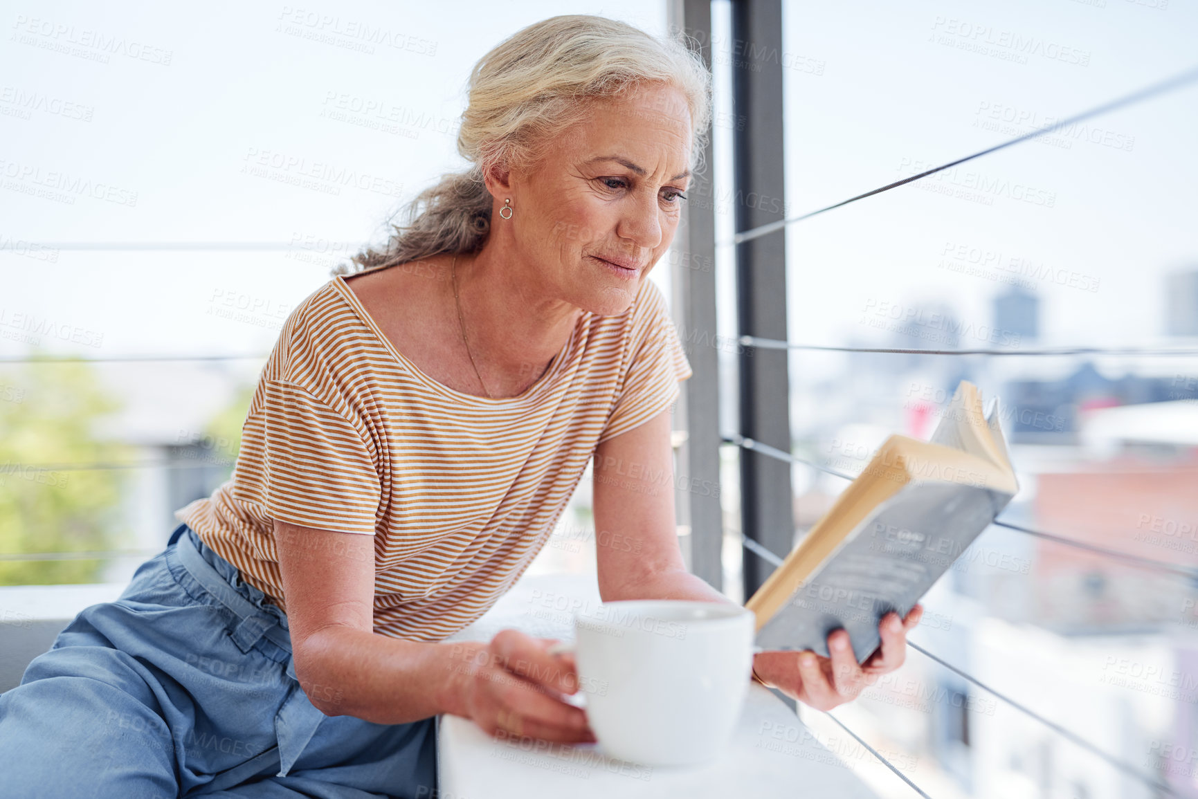 Buy stock photo Cropped shot of an attractive senior woman enjoying a cup of coffee while reading a book outside on her balcony at home