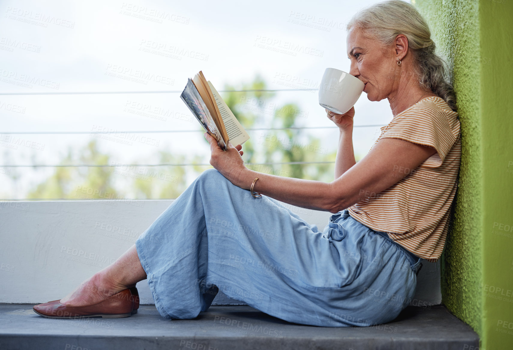 Buy stock photo Full length shot of an attractive senior woman enjoying a cup of coffee while reading a book outside on her balcony at home