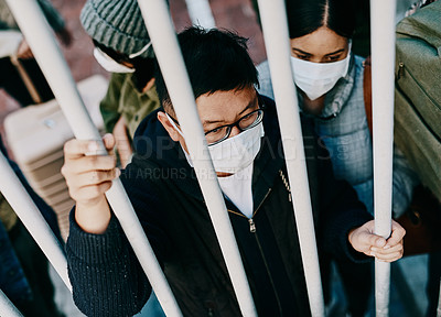Buy stock photo Shot of a young man wearing a mask while stuck behind a gate in a foreign city