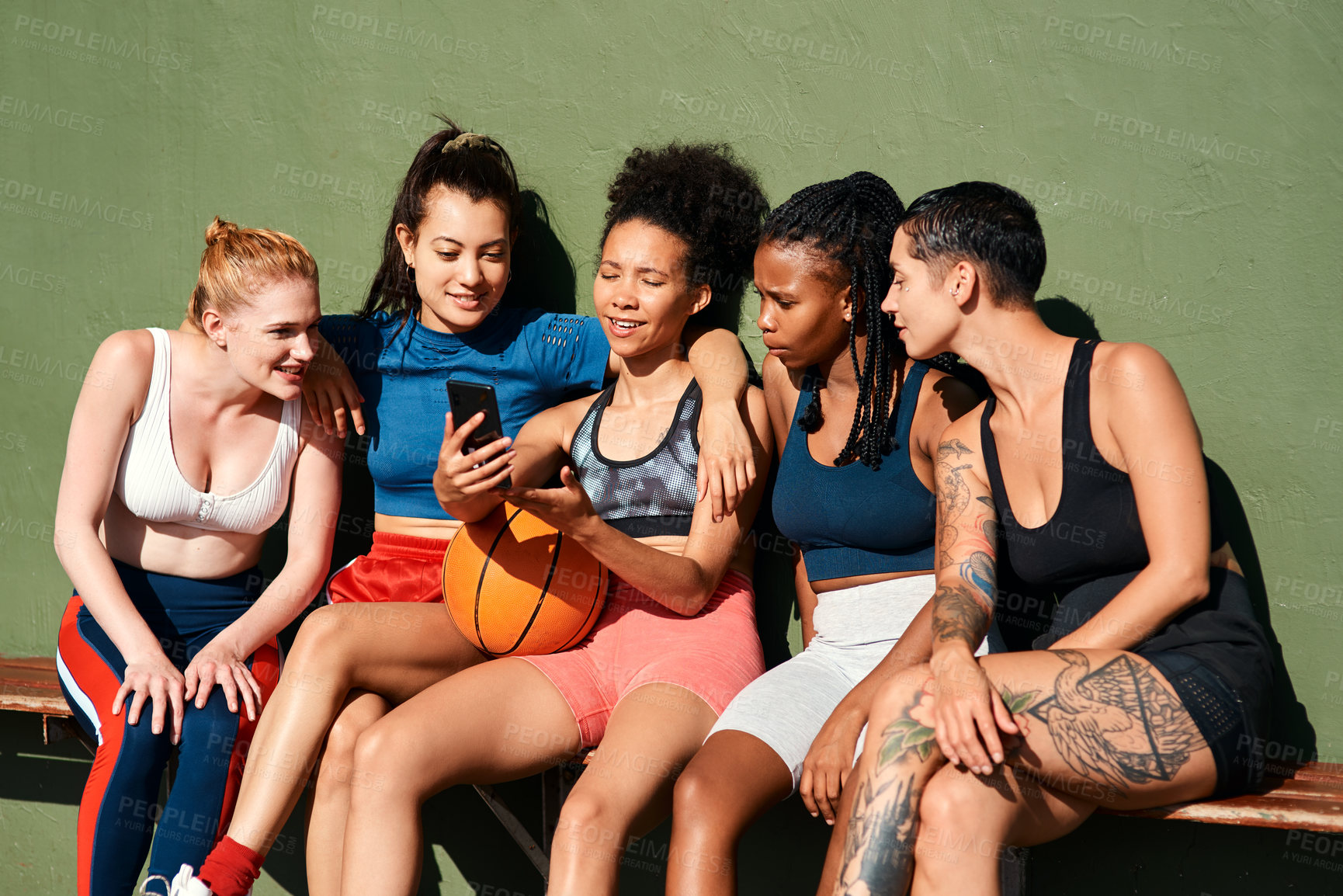 Buy stock photo Cropped shot of a diverse group of sportswomen sitting together after playing basketball and looking at a cellphone