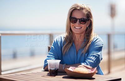 Buy stock photo Portrait of a carefree middle aged woman having lunch outside next to a beach promenade during the day