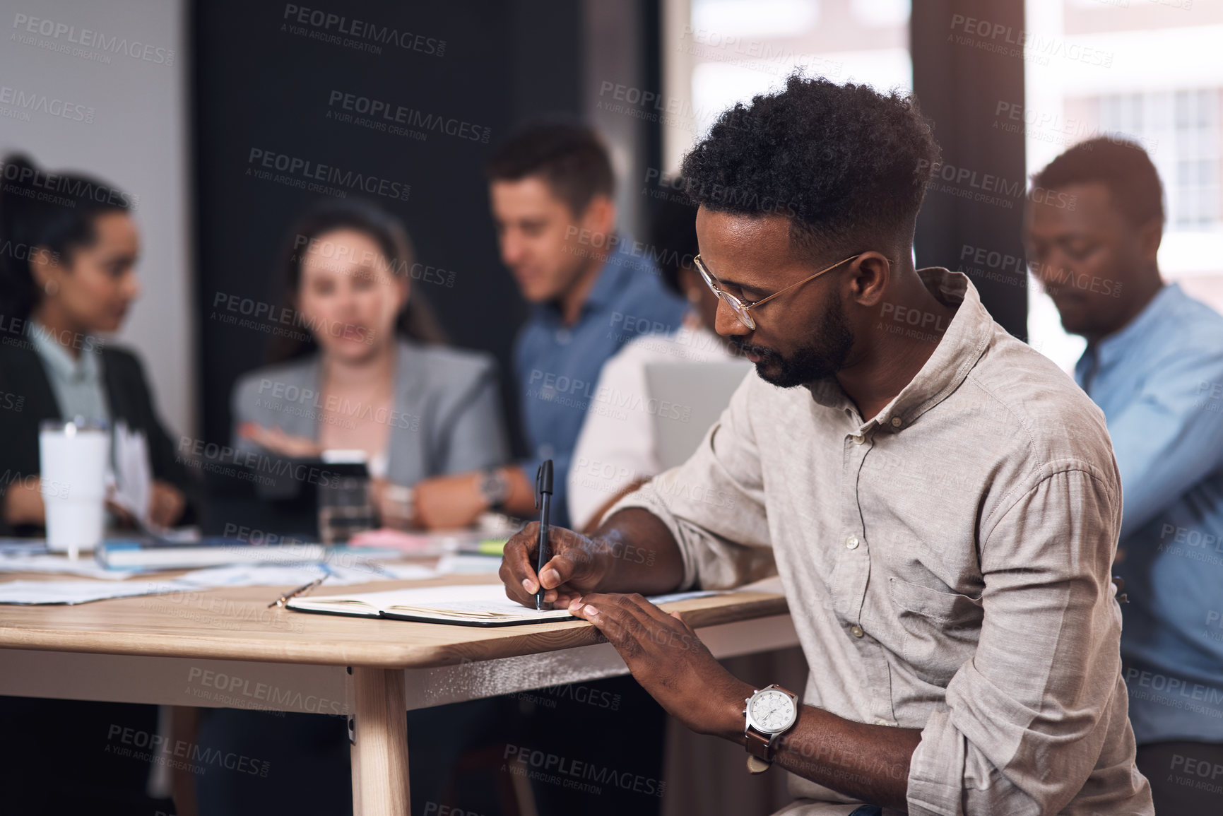 Buy stock photo Shot of a young businessman writing in a notebook while sitting in an office with his colleagues in the background