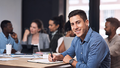 Buy stock photo Portrait of a young businessman writing in a notebook while sitting in an office with his colleagues in the background