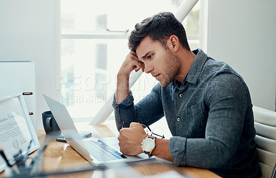 Buy stock photo Cropped shot of a handsome young businessman sitting alone and feeling stressed while using his laptop in the office