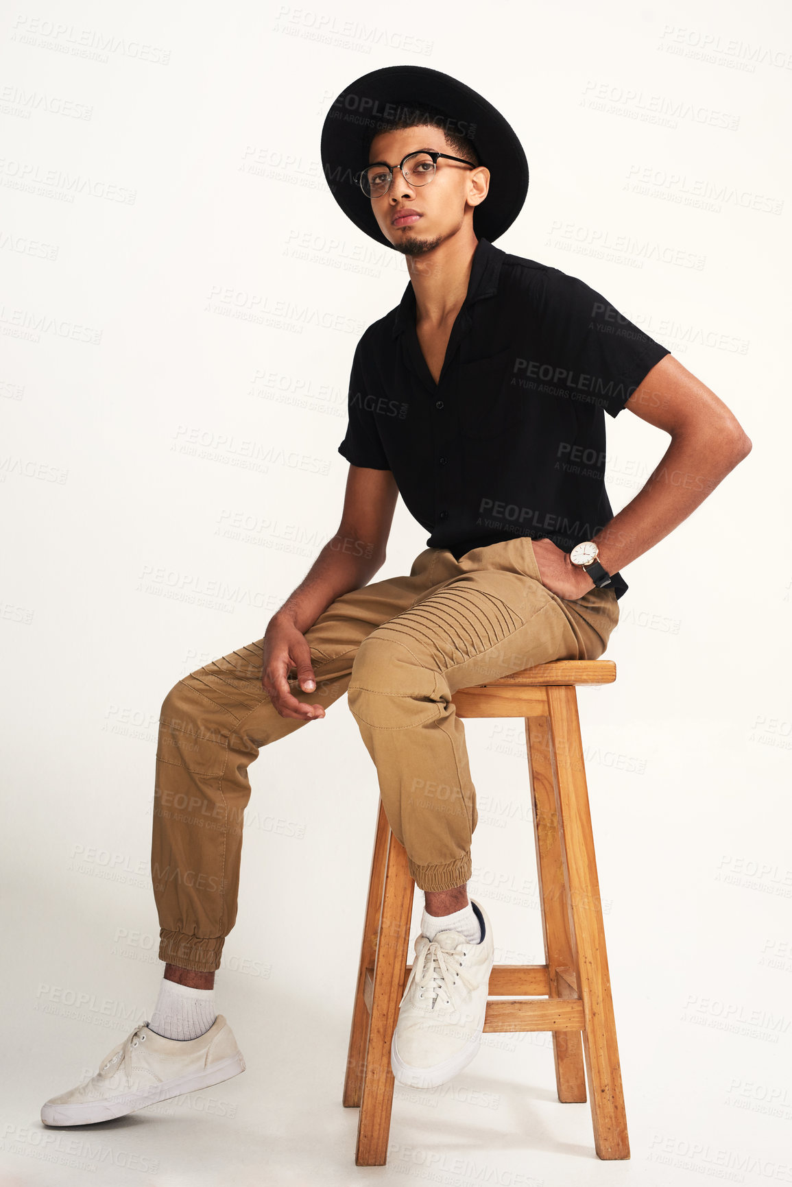 Buy stock photo Portrait of a handsome young man wearing a hat and glasses while being seated on a chair against a grey background inside of a studio