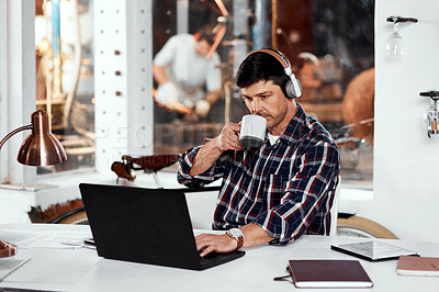 Buy stock photo Cropped shot of a handsome young businessman drinking coffee while working on a laptop in an office inside his workshop