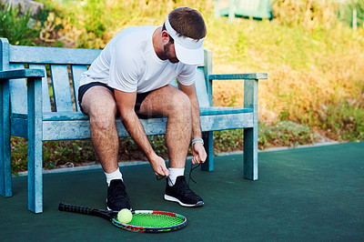 Buy stock photo Shot of a sporty young man tying his laces while sitting on a bench on a tennis court
