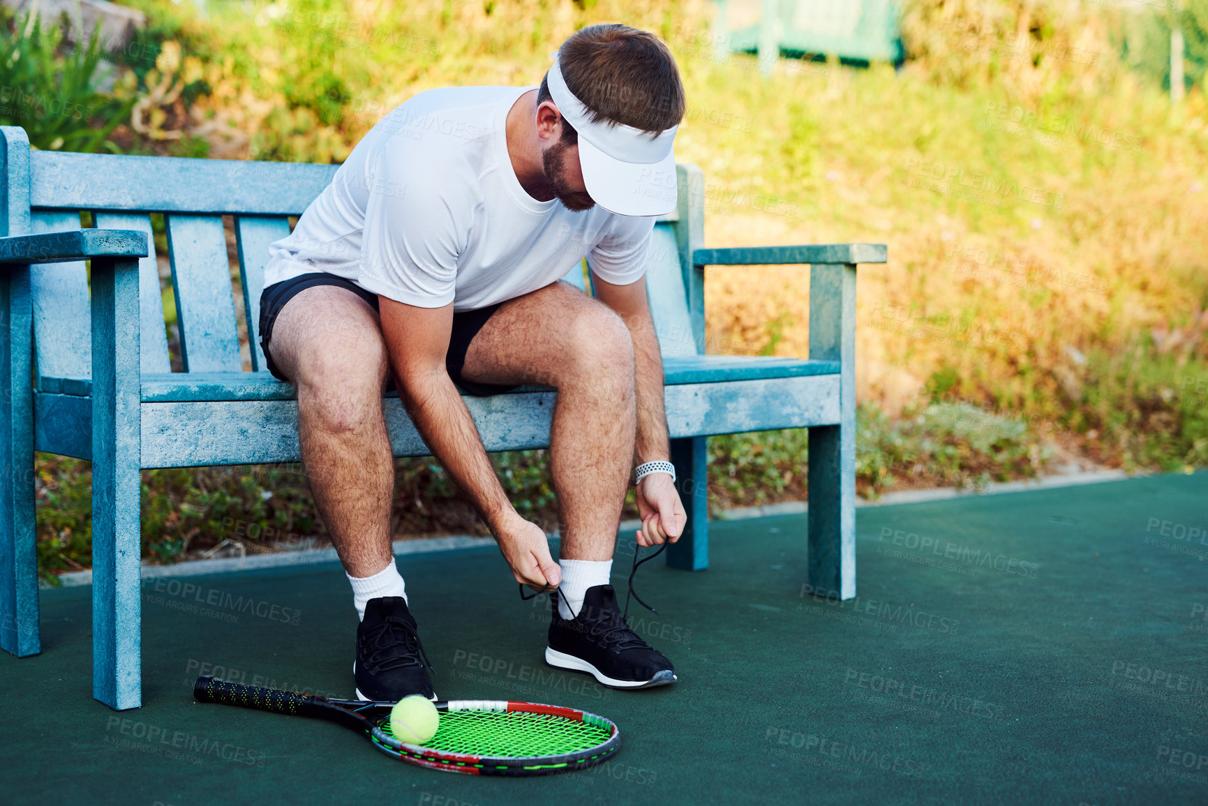 Buy stock photo Shot of a sporty young man tying his laces while sitting on a bench on a tennis court