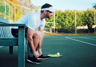 Buy stock photo Shot of a sporty young man tying his laces while sitting on a bench on a tennis court