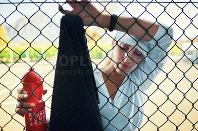 Buy stock photo Shot of a sporty young woman looking exhausted while leaning against a fence on a tennis court