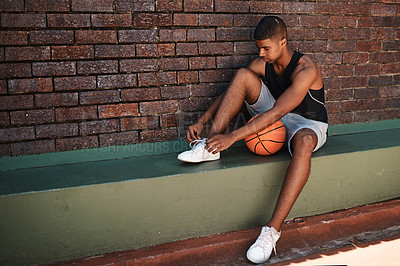 Buy stock photo Shot of a sporty young man tying his laces while playing basketball outdoors