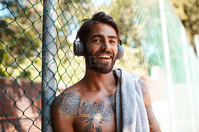 Buy stock photo Portrait of a sporty young man listening to music while standing against a fence outdoors