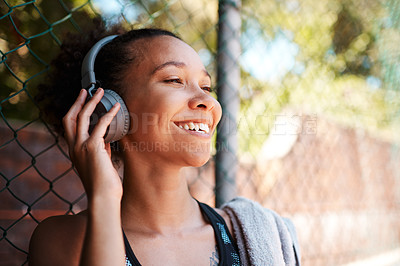Buy stock photo Shot of a sporty young woman listening to music while standing against a fence outdoors