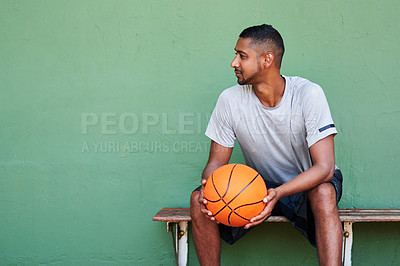 Buy stock photo Shot of a sporty young man holding a basketball while sitting on a bench against a wall