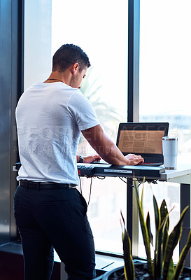 Buy stock photo Shot of a young businessman working on a laptop while walking on a treadmill in an office