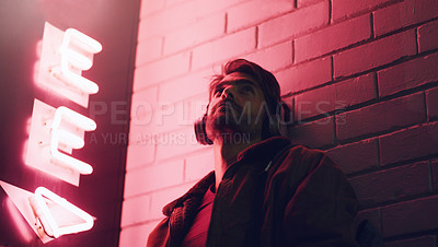 Buy stock photo Shot of a young man wearing headphones while leaning against a building at nighttime