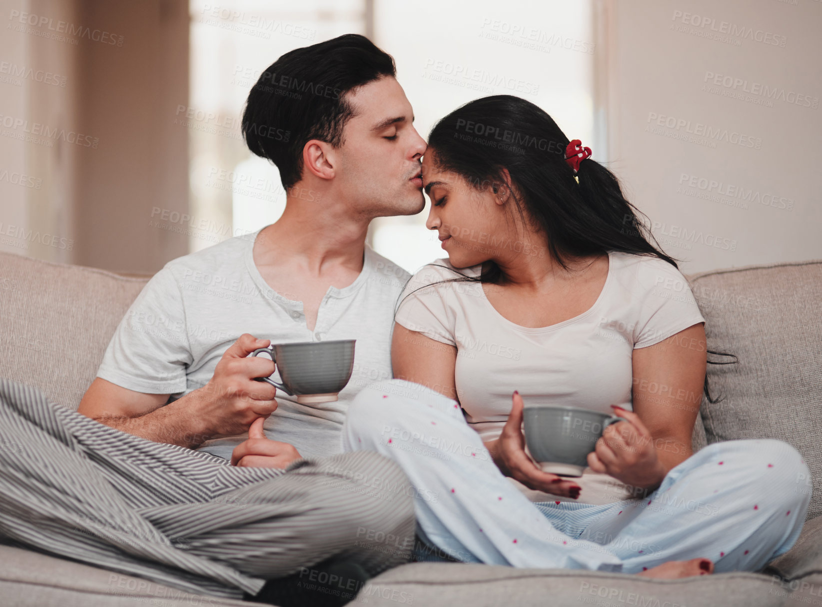 Buy stock photo Cropped shot of a handsome young man kissing his wife on the forehead while sitting on the living room sofa