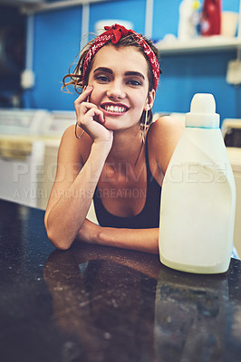 Buy stock photo Portrait of an attractive young woman resting her arms on a counter with a bottle of bleach next to her while waiting for her washing to wash inside of a laundry room