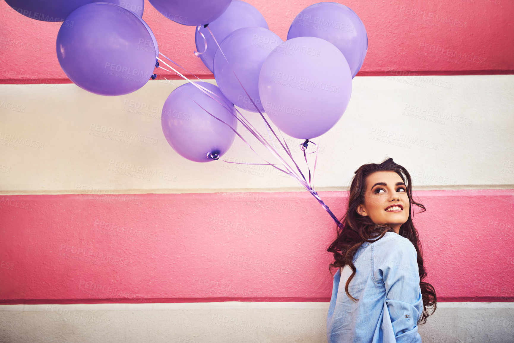 Buy stock photo Cropped shot of a beautiful young woman holding purple balloons against a wall outside