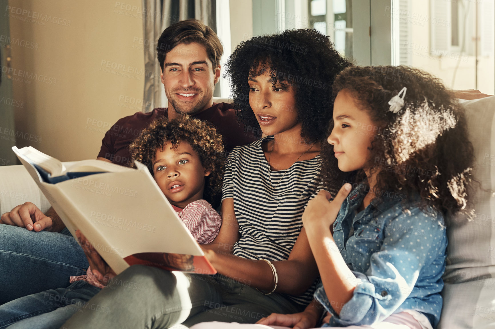 Buy stock photo Shot of a beautiful young family of four reading a story book together while relaxing on a sofa at home