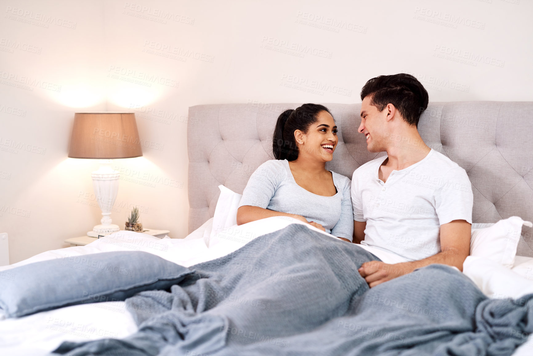 Buy stock photo Bedroom, love and interracial couple laughing together in home holding hands at night. Smiling, diversity and marriage of an Indian woman and man with care and relax happiness with a smile in a house