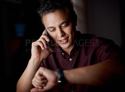 Buy stock photo Shot of a young businessman checking the time on his watch while talking on a cellphone in an office at night