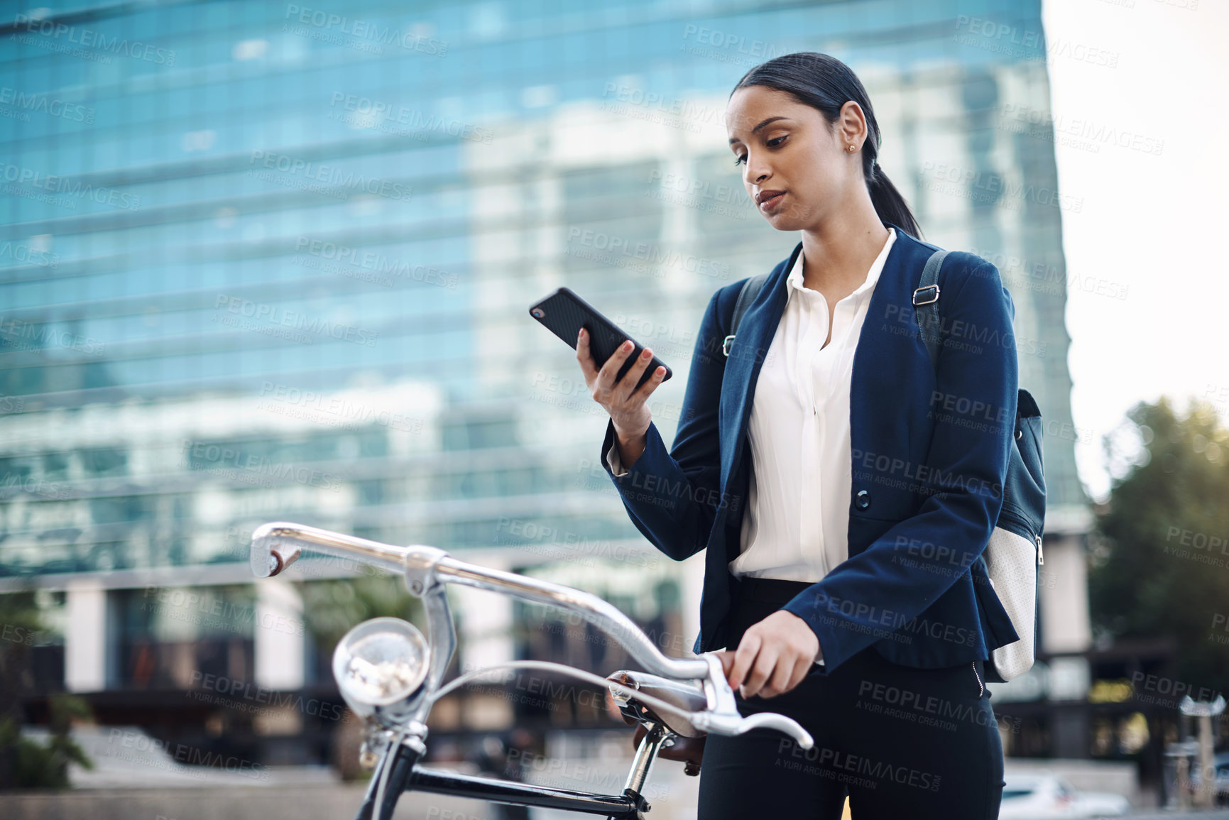 Buy stock photo Shot of a young businesswoman using a smartphone while traveling with a bicycle through the city