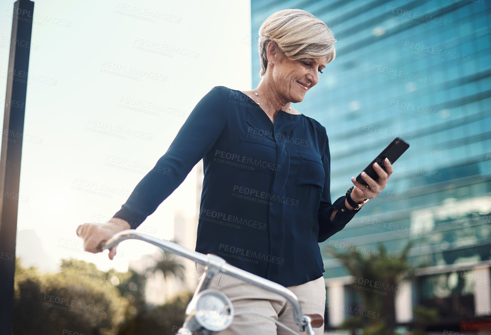 Buy stock photo Shot of a mature businesswoman using a smartphone while traveling with a bicycle through the city