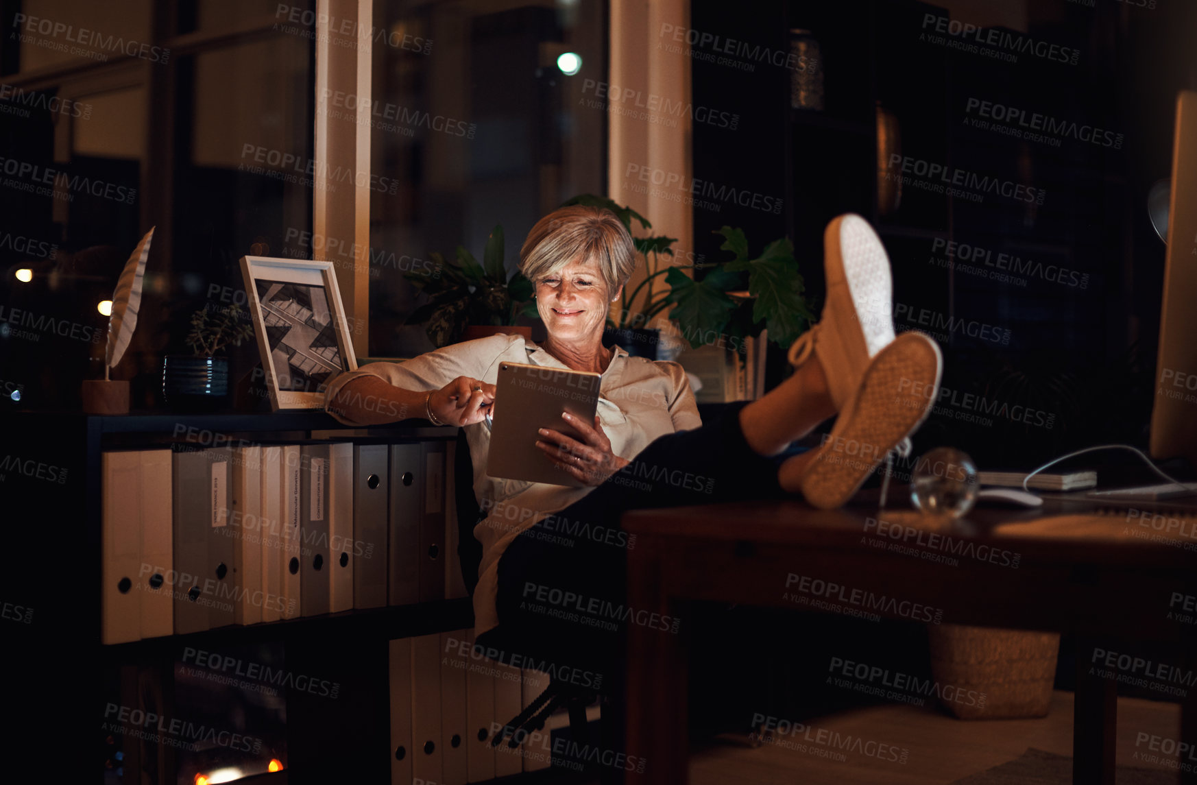 Buy stock photo Full length shot of an attractive mature businesswoman looking relaxed while using her tablet late at night in the office