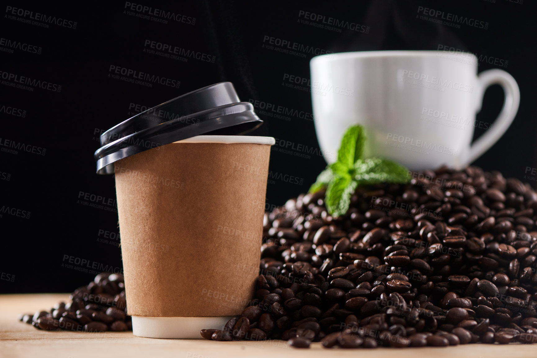 Buy stock photo Closeup shot of a paper cup on a surface, with a teacup and mint leaf resting on a pile of coffee beans in the background