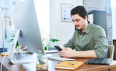 Buy stock photo Shot of a young businessman using a cellphone while working on a computer in an office
