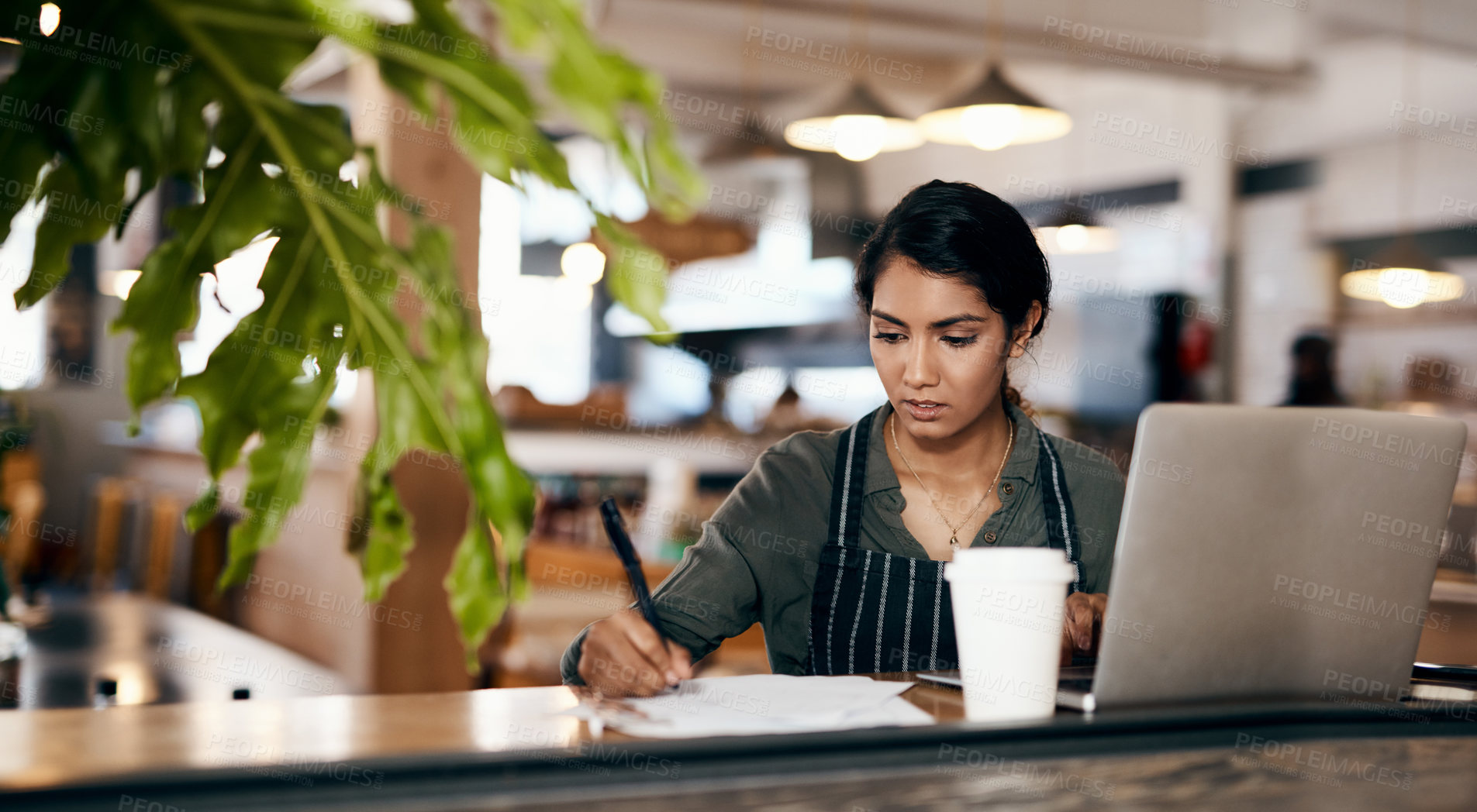 Buy stock photo Shot of a young woman using a laptop and going over paperwork while working in a cafe