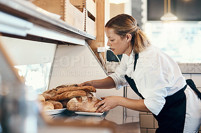 Buy stock photo Shot of a mature woman putting a selection of freshly baked breads on display in her bakery