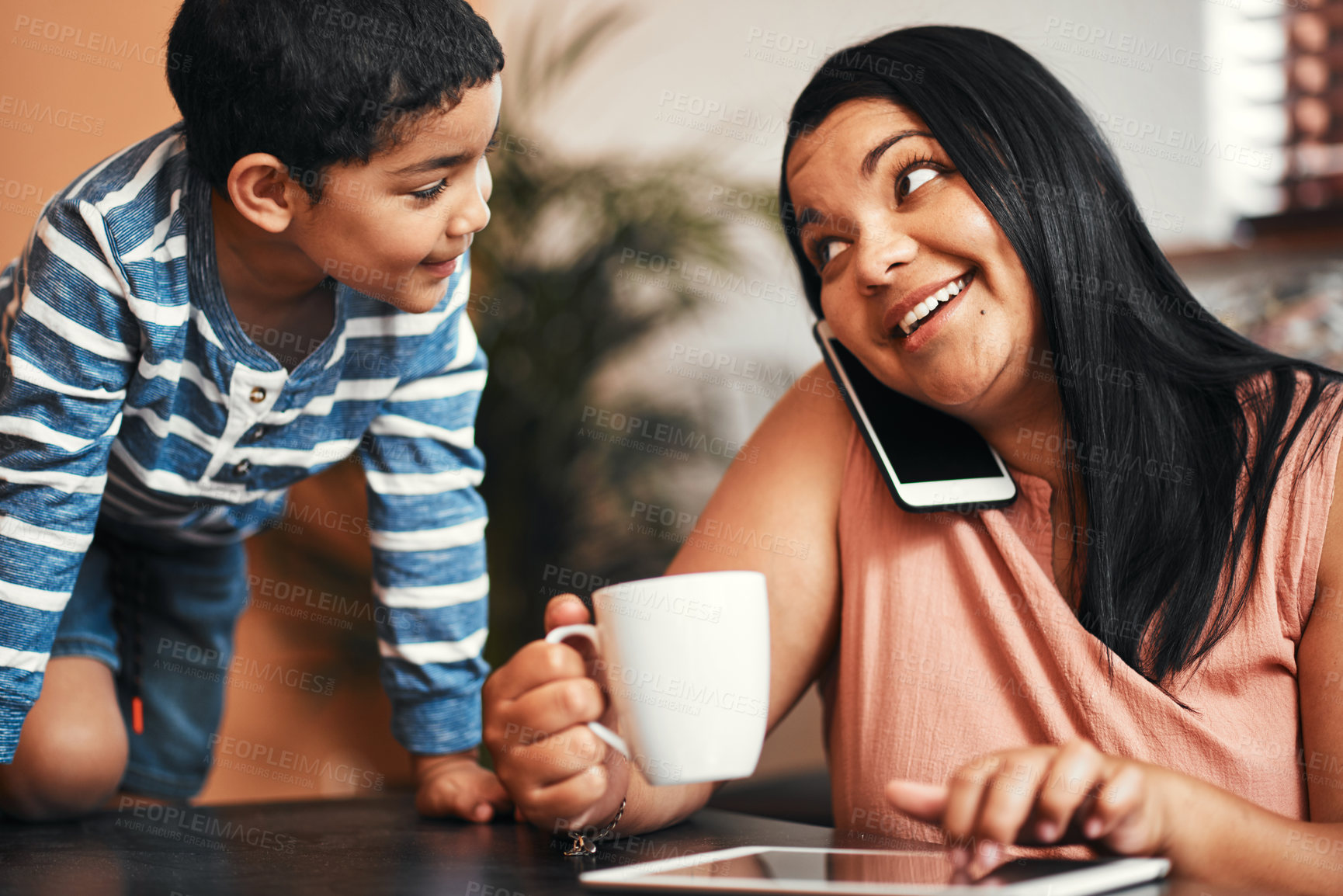 Buy stock photo Shot of a mother talking on a cellphone while using a digital tablet with her son alongside her at home