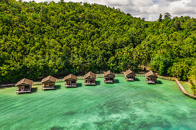 Buy stock photo Aerial, bungalow and resort by tropical ocean with hill, island and trees on beach for tourism agency. Jetty, hotel and hospitality by sea, adventure and drone in nature with water in Raja Ampat