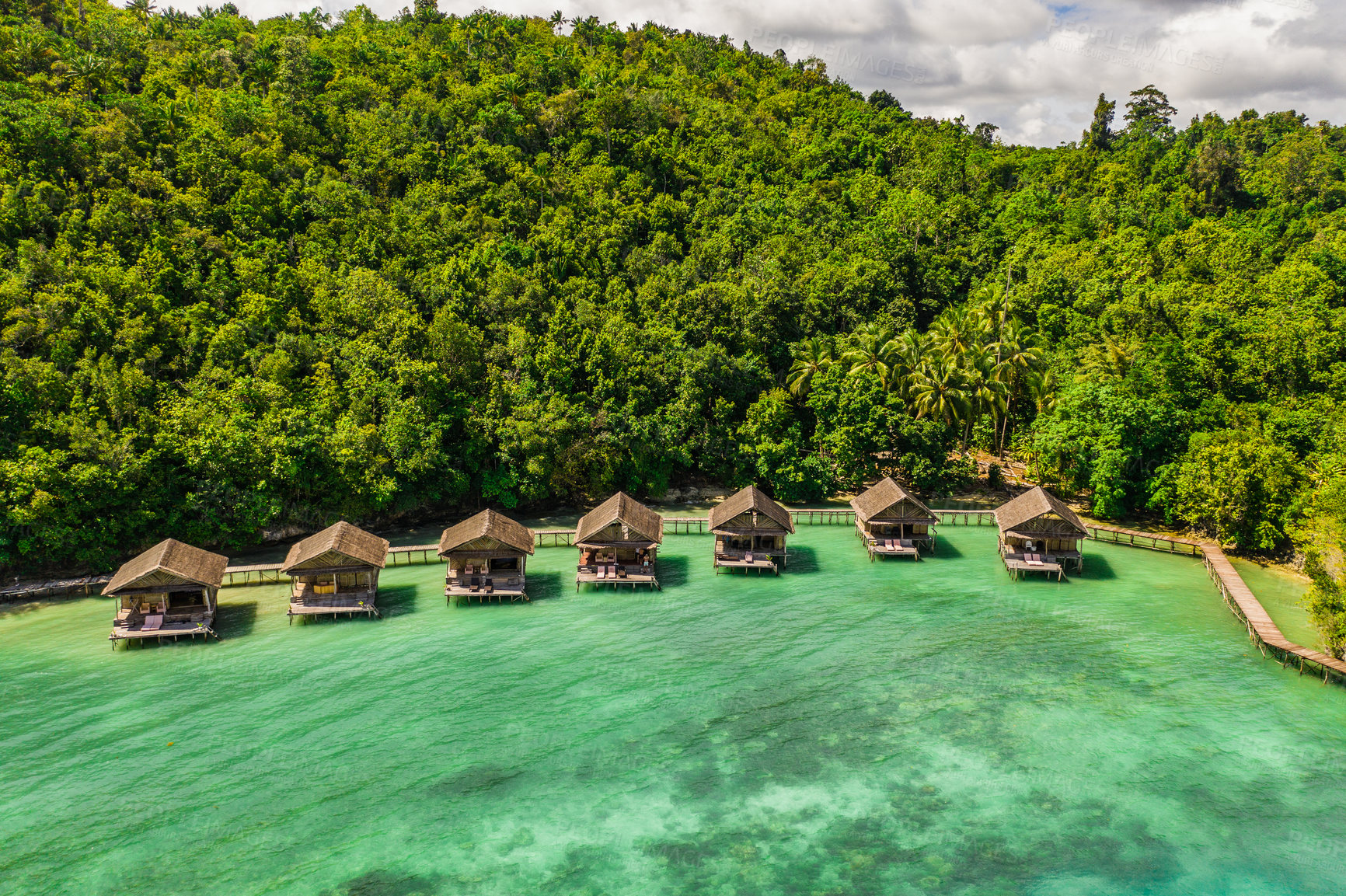 Buy stock photo Aerial, bungalow and resort by tropical ocean with hill, island and trees on beach for tourism agency. Jetty, hotel and hospitality by sea, adventure and drone in nature with water in Raja Ampat