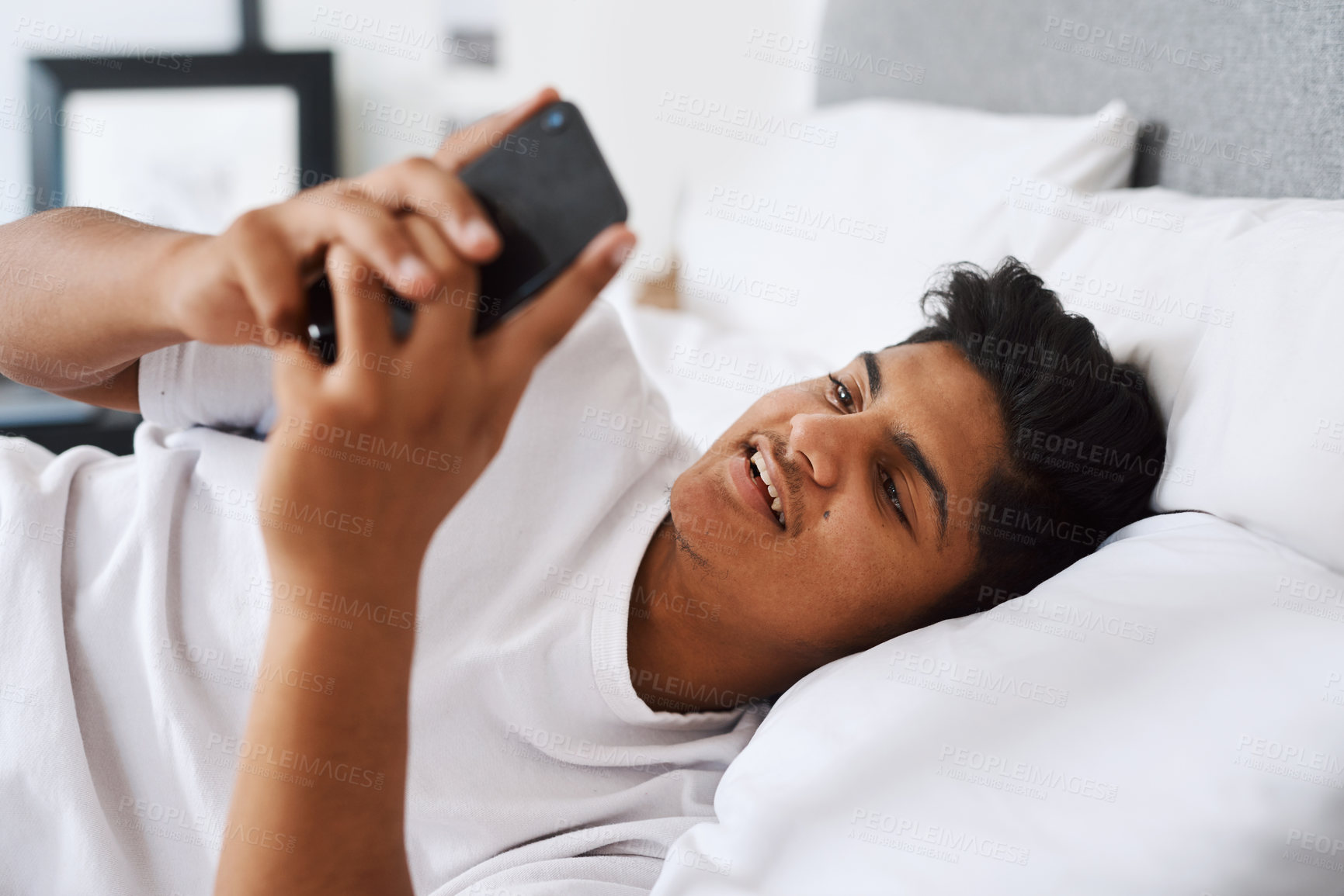 Buy stock photo Shot of a young man using his cellphone while lying on his bed