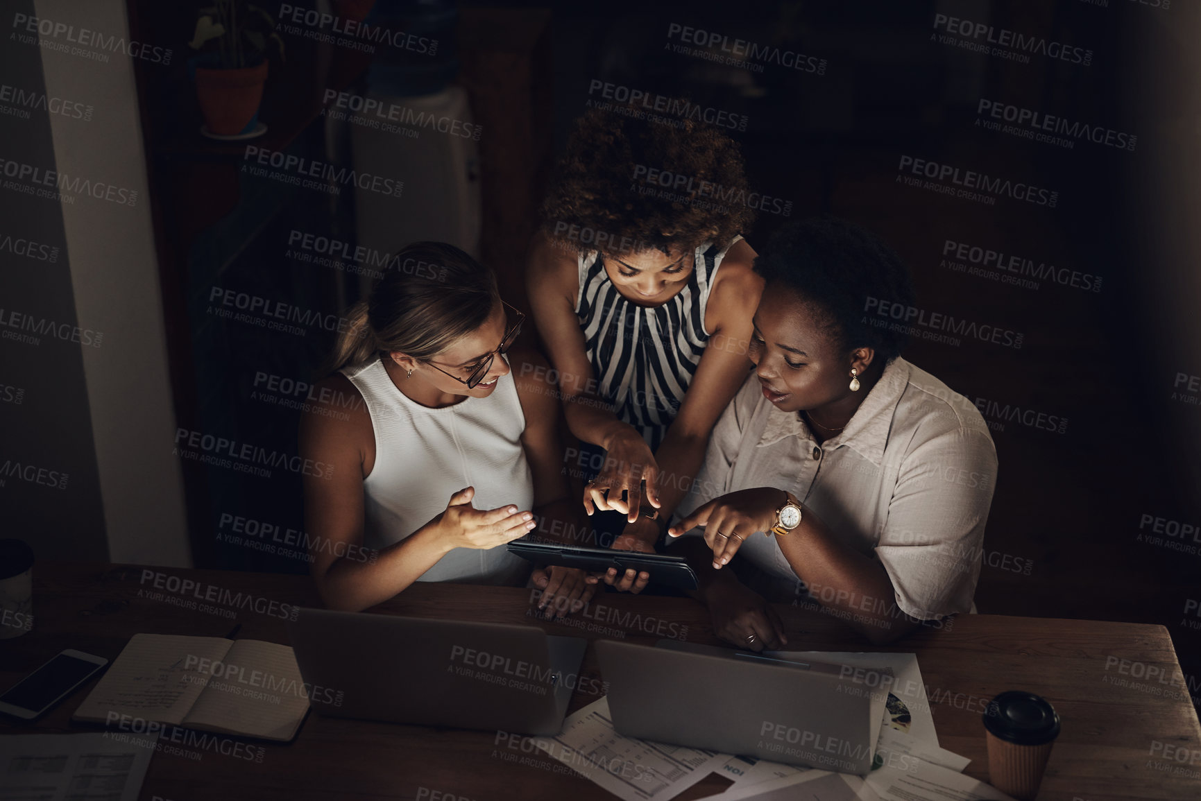 Buy stock photo Shot of a group of young businesswomen using a laptop and digital tablet during a late night at work