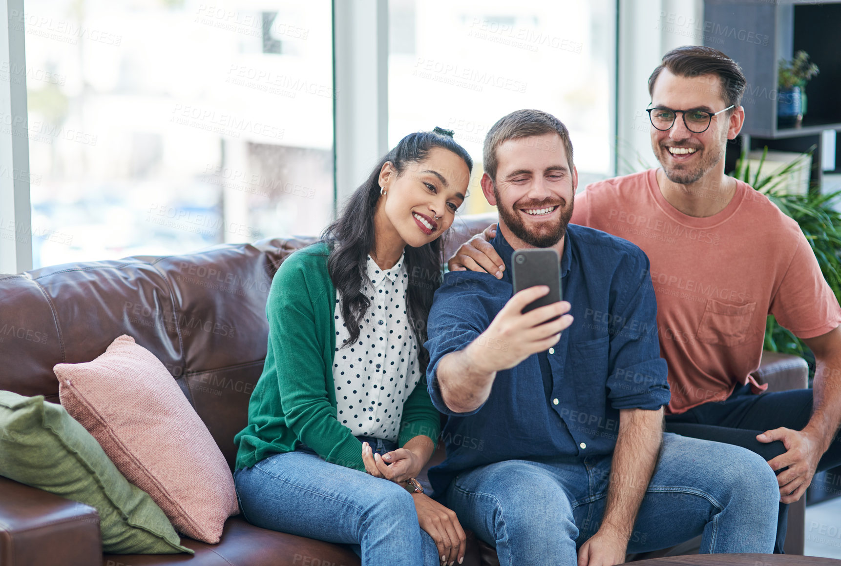 Buy stock photo Shot of two young men and a woman taking selfies together on a sofa
