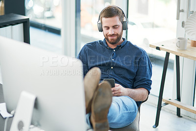 Buy stock photo Shot of a young businessman using a smartphone and headphones in a modern office