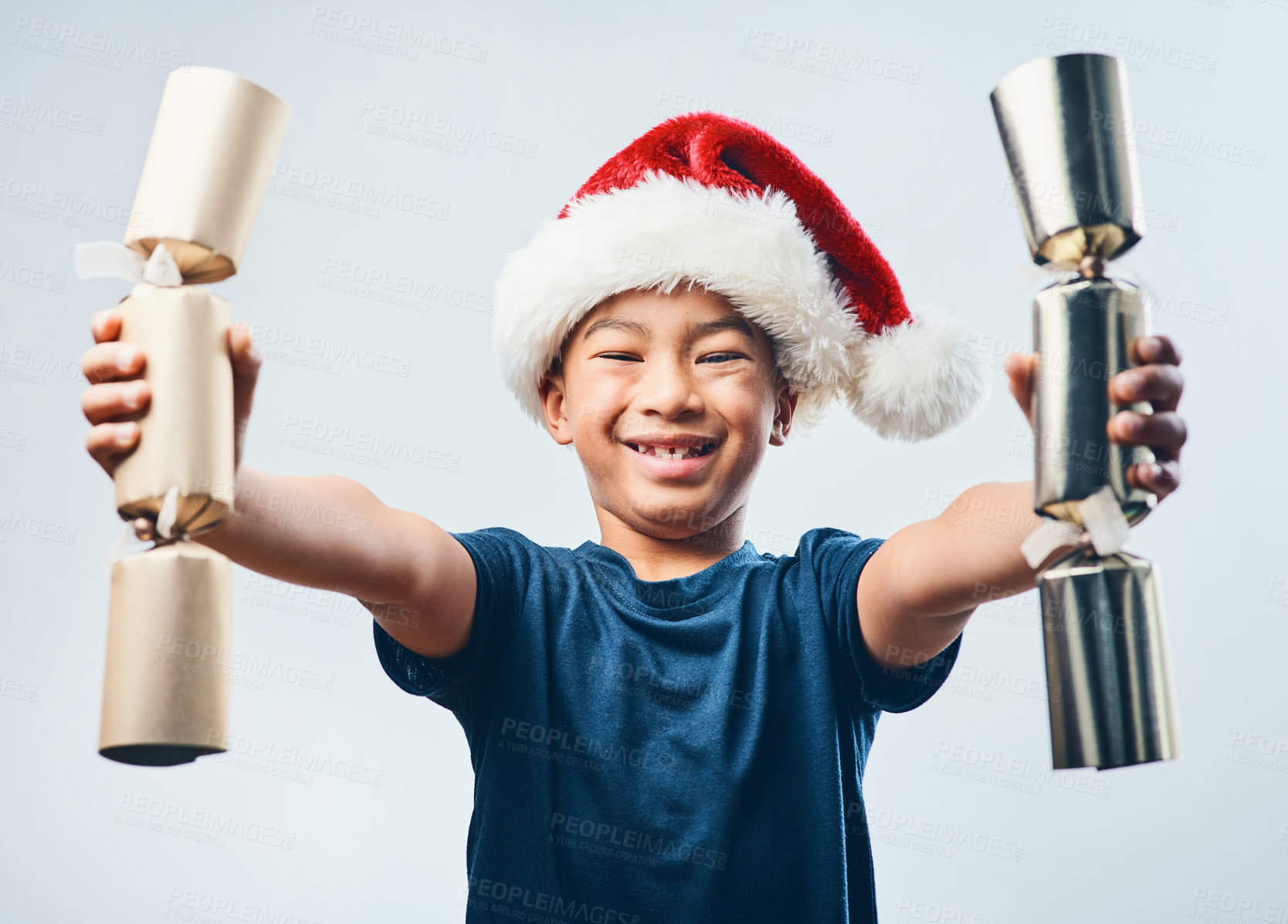 Buy stock photo Studio shot of a cute little boy wearing a Santa hat and holding two Christmas crackers against a grey background