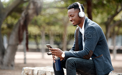 Buy stock photo Businessman browsing social media on his phone and enjoying his music playlist app in headphones outdoors. Young, happy and casual african male relaxing in a nature park outside during a break 