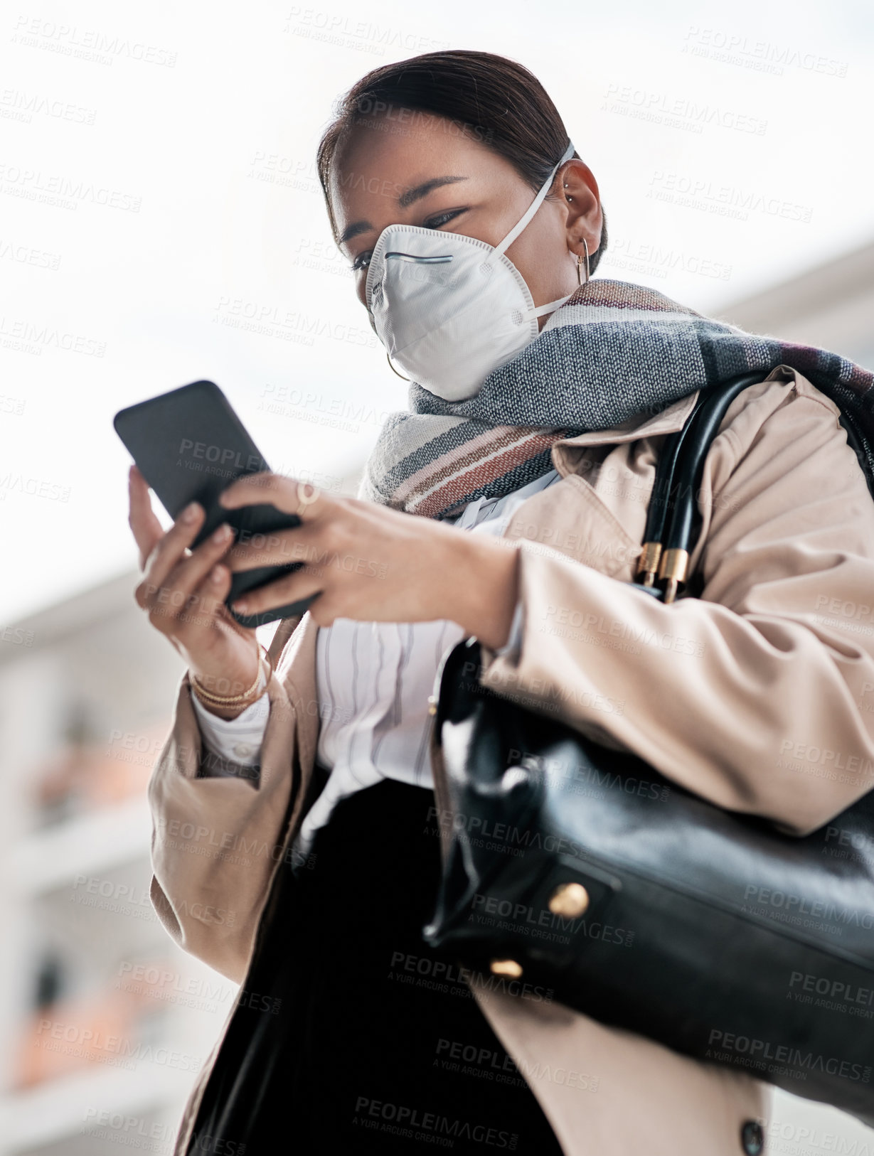 Buy stock photo Shot of a young woman wearing a mask and using a smartphone while out in the city